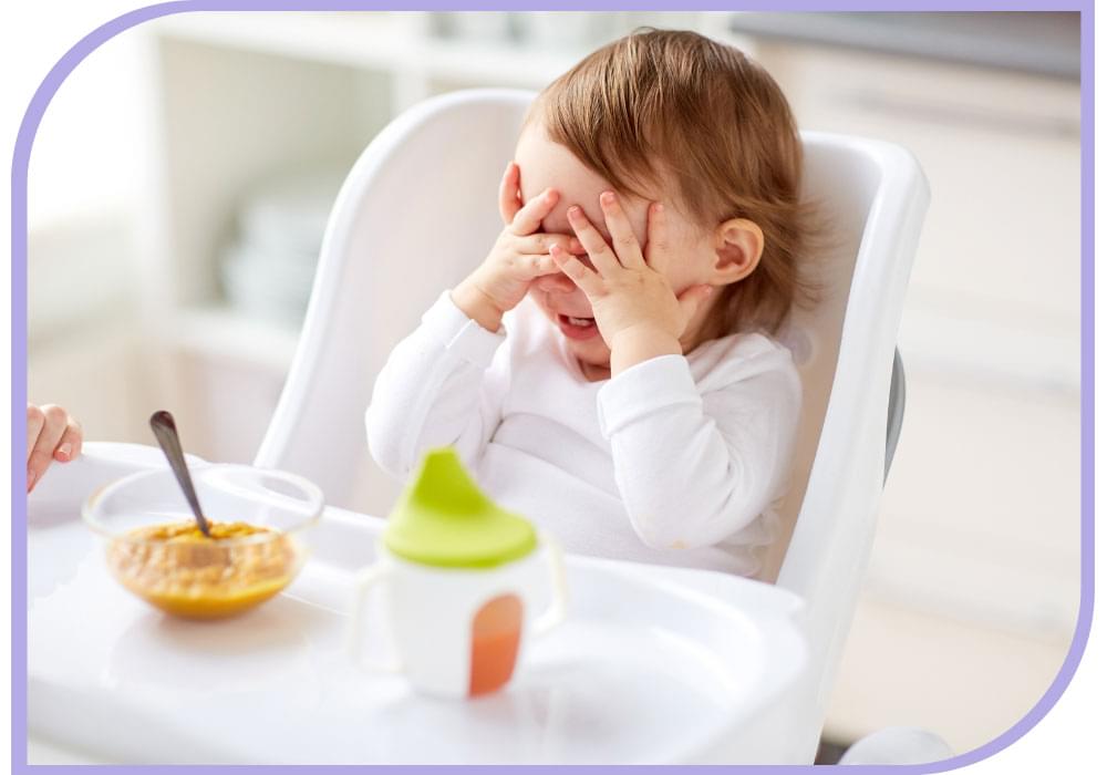 Young child with white shirt in a white high chair has a bowl of orange coloured puree with a spoon and a white sippy cup with a green lid in front of them. Child has both hands covering there eyes/face. Feeding Therapy | Able Kids Therapy