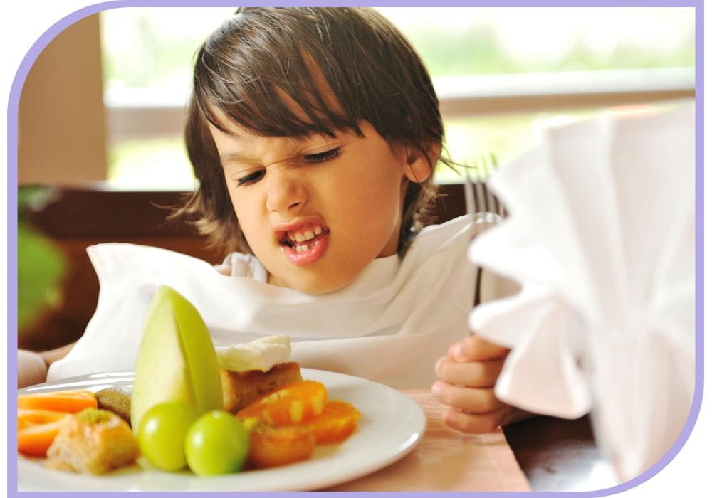 Young child sitting at the table with a white napkin covering their clothes. Has a plate of assorted fruit in front of them. The child is holding a silver fork in their left hand while looking down at the plate of fruit with a screwed up face. Feeding Therapy | Able Kids Therapy