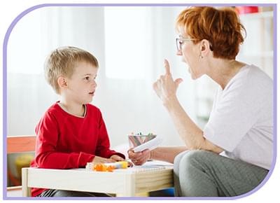 Able Kids Therapy Allied Health Services - Speech Therapy & Paediatric Occupational Therapy. Child sits at a low table with a therapist who is helping the child hear/understand the sounds her mouth is making while encouraging him to copy.