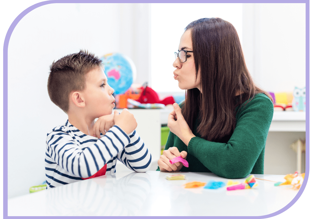 A young child and a Speech therapy specialist sit at a white table with some colourful objects on it. The specialist is holding a small pink deflated balloon in her right hand resting it on the table. The Specialist is puckering her lips together, the young boy is looking at her lips and copying it.