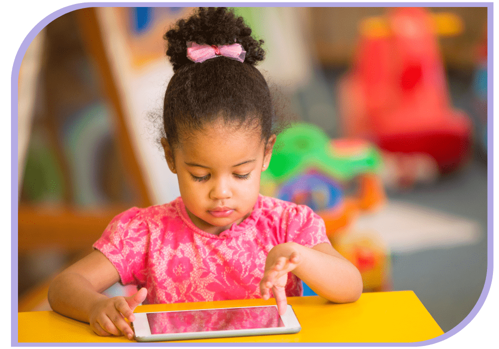 A young girl in a pink dress is sitting at a yellow table with a phone laying horizontal on the table in front of her. She is looking down at the phone with her left thumb pressing down onto the phone. Speech Therapy | Able Kids Therapy