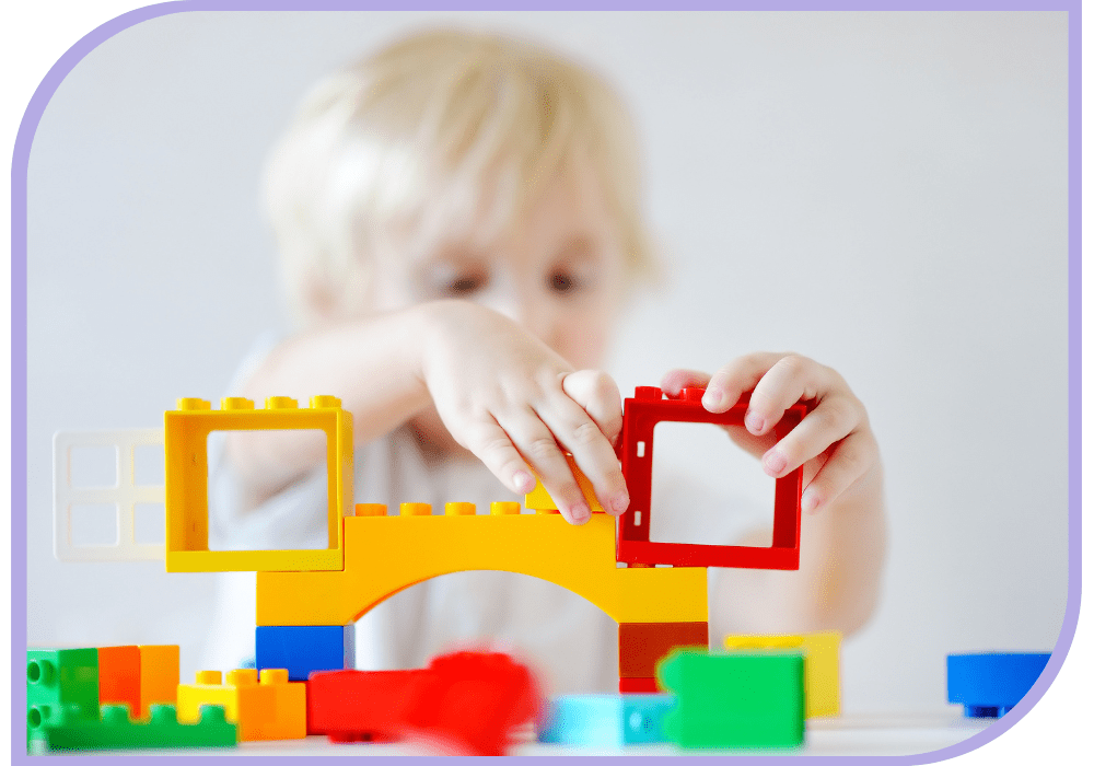 A young child dressed in white sitting at a table with colourful building blocks. The child is using both hands to build a house. Group Therapy | Able Kids Therapy